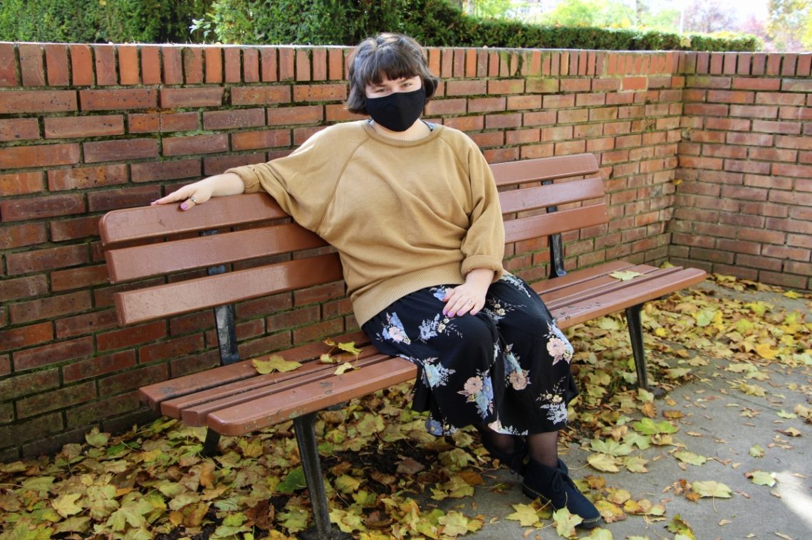 Maria sits on a brown bench. The ground is littered with yellow leaves.