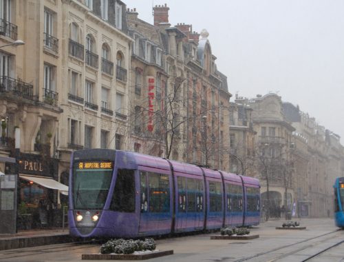 One purple and one blue tram on a snowy day on a street in Reims, France.