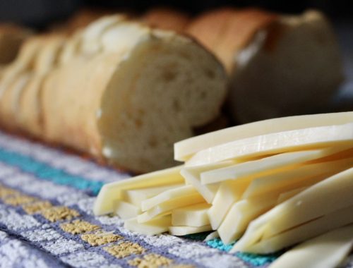 Two baguettes and a pile of cheese slices sit on top of a purple, white, blue, and yellow kitchen towel.