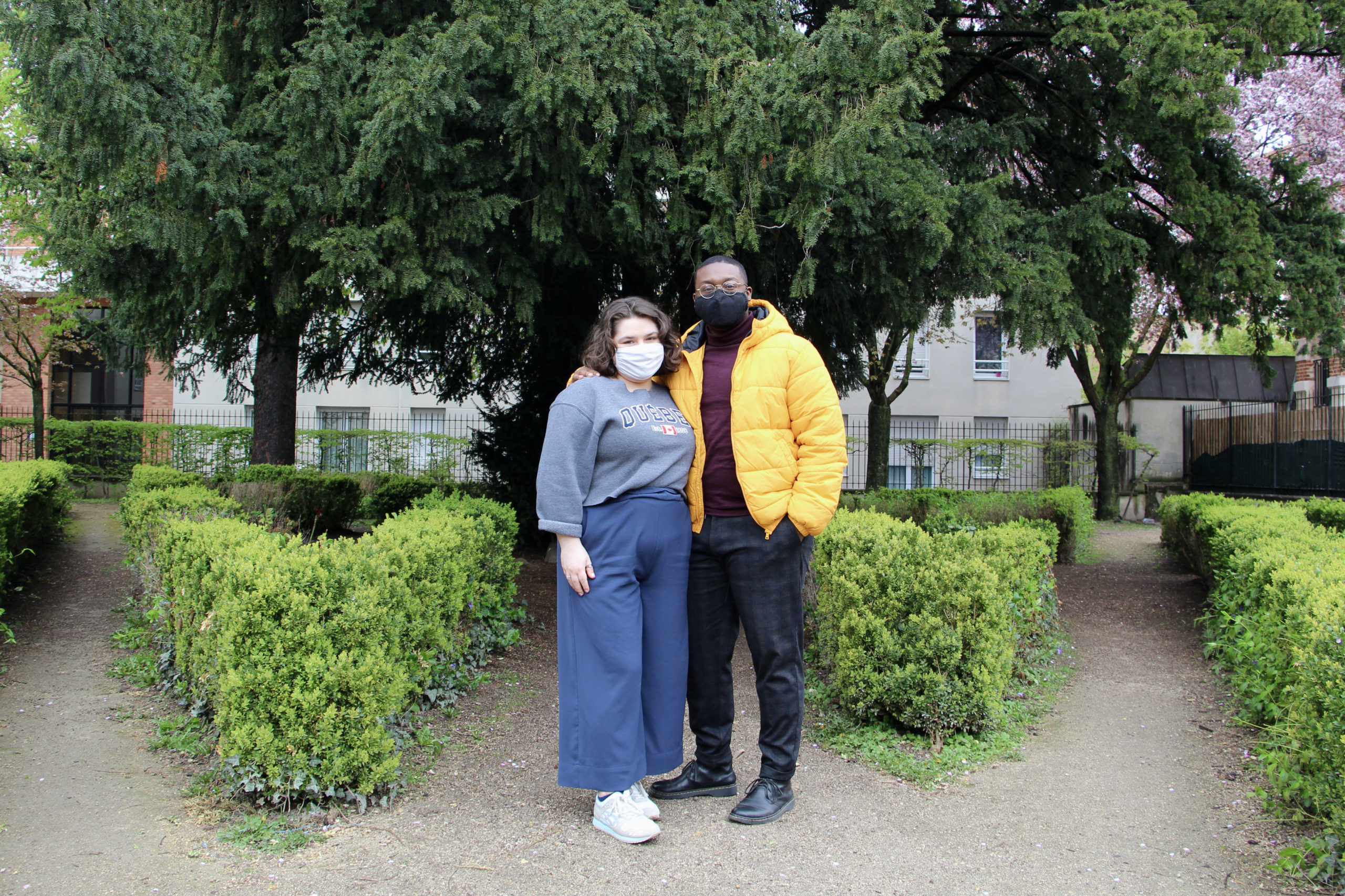 Maria and Jalen stand side by side in a park in Reims, France with green bushes and trees in the background.