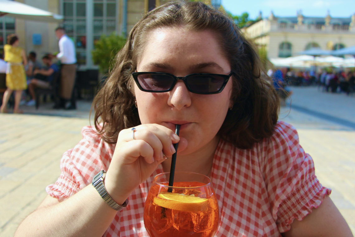 Maria sips an Apérol Spritz at a French café in a gingham dress and sunglasses..