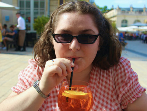 Maria sips an Apérol Spritz at a French café in a gingham dress and sunglasses..