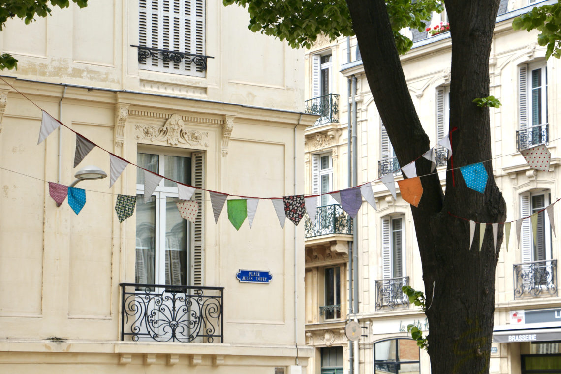 Colorful banners strung through a tree in front of beige buildings in Reims, France.