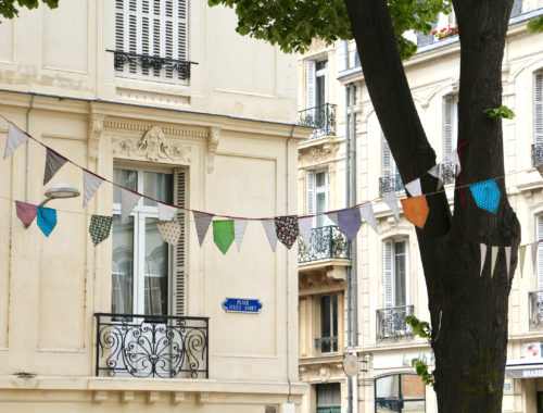 Colorful banners strung through a tree in front of beige buildings in Reims, France.