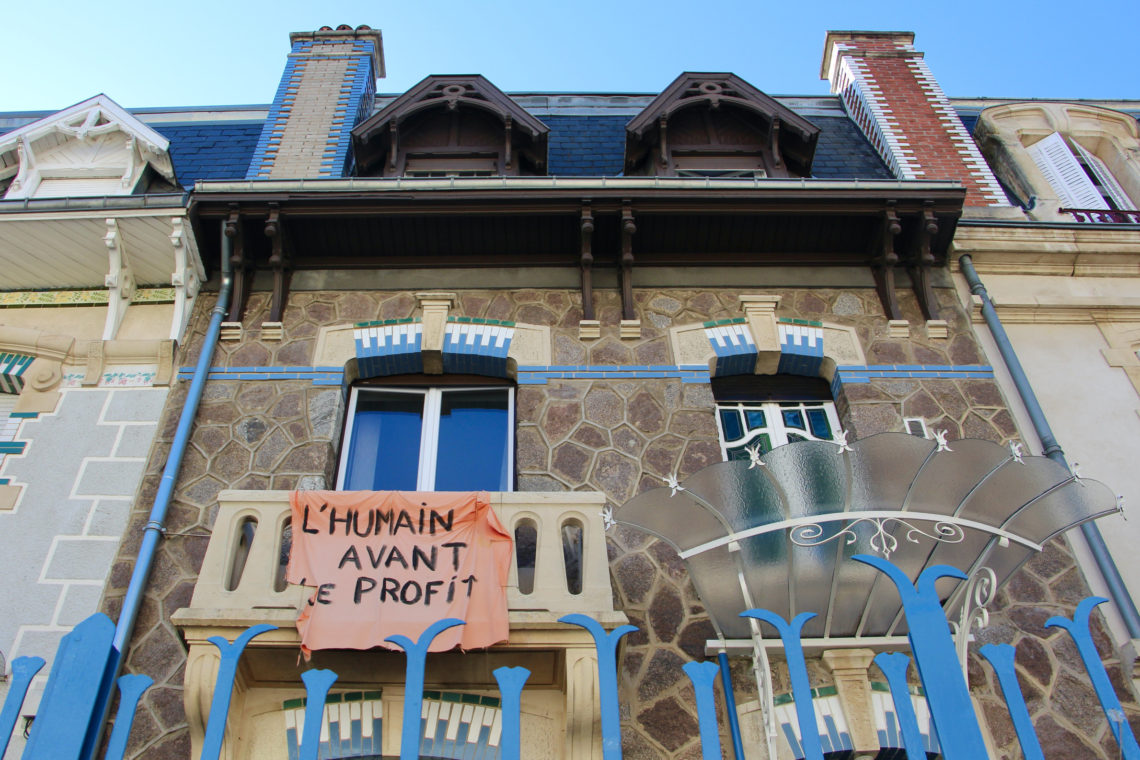 Art deco buildings in Nancy, France display a tattered fabric sign that reads L'HUMAIN AVANT LE PROFIT.