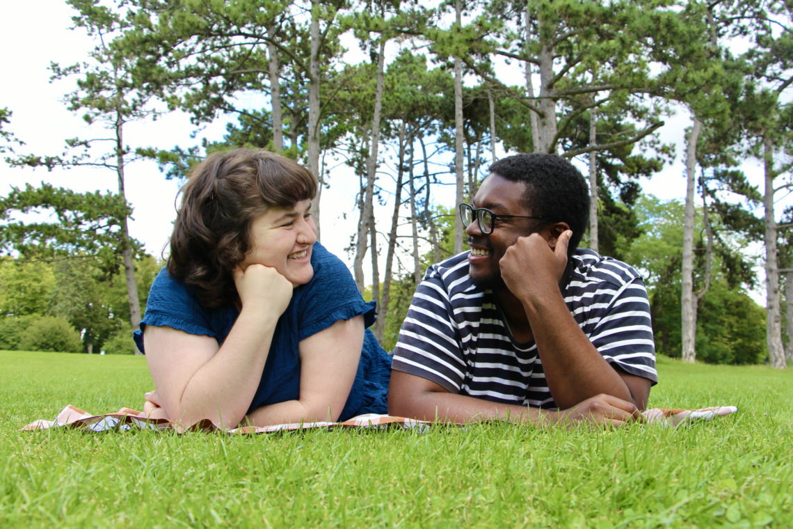 Maria and Jalen lay on a picnic blanket in the grass and smile at each other in Reims, France.