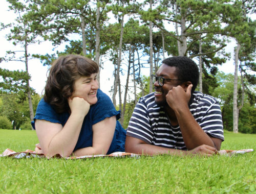 Maria and Jalen lay on a picnic blanket in the grass and smile at each other in Reims, France.
