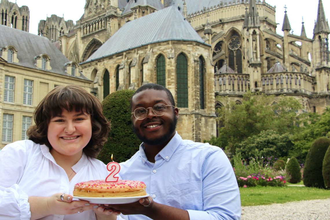 Jalen and Maria pose with a cake behind the Cathédrale de Reims.