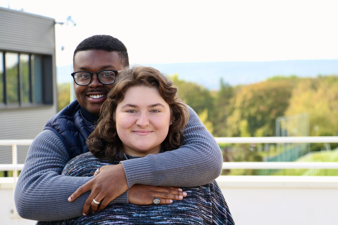 Jalen hugs Maria on the URCA campus with a bright sky and foliage in the background.