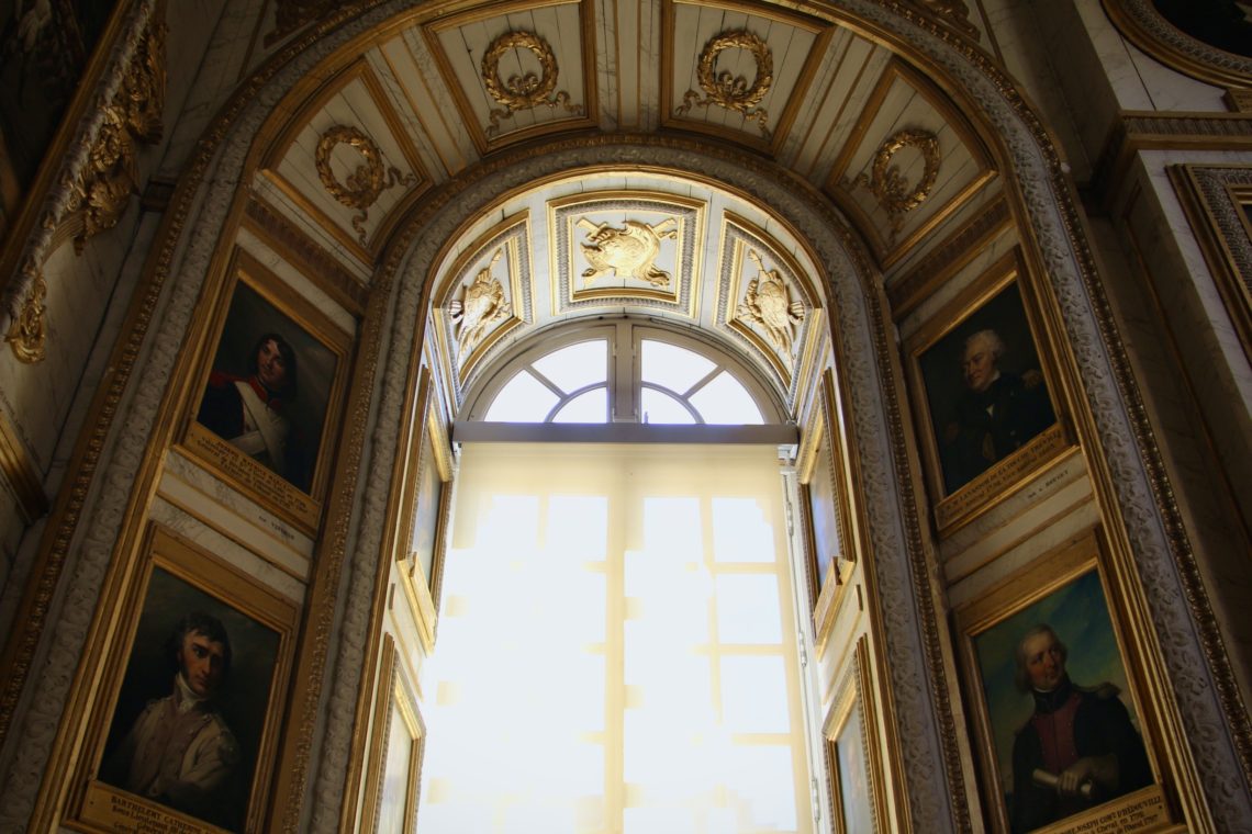 Sunlight pours through a window in the middle of an archway decorated with portraits in the Palace of Versailles.
