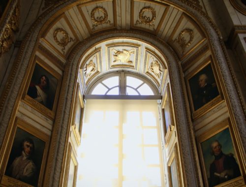 Sunlight pours through a window in the middle of an archway decorated with portraits in the Palace of Versailles.