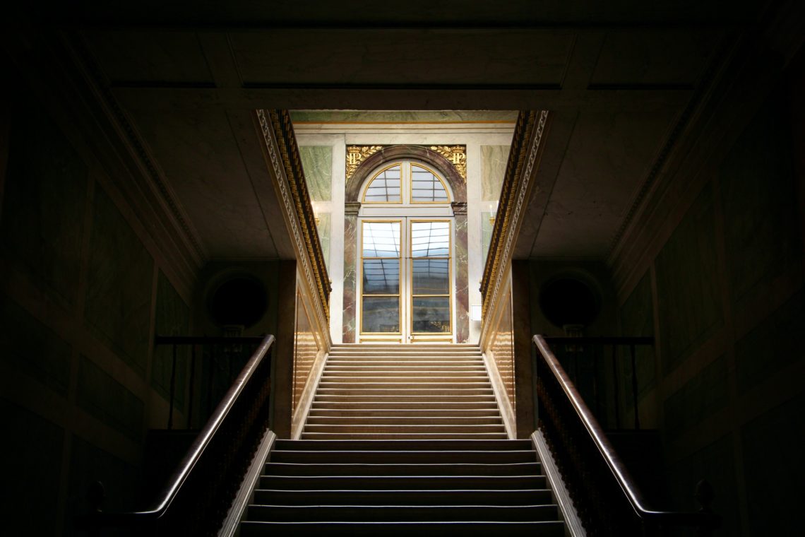 A bright stairwell contrasts with a dark room in the Palace of Versailles.