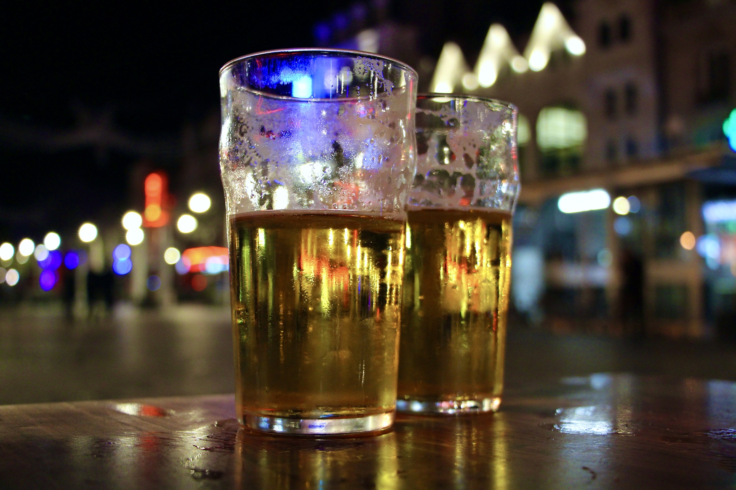 Two pints of beer on a table outside illuminated by city lights in the evening.